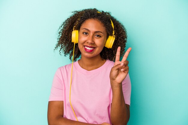 Joven afroamericana escuchando música aislada sobre fondo azul mostrando el número dos con los dedos.