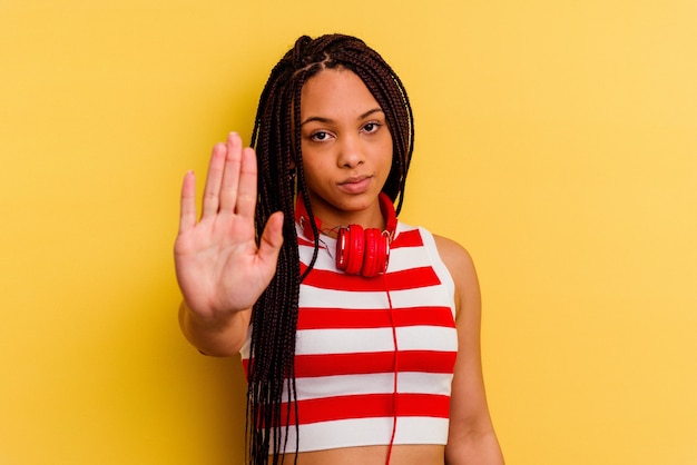 Joven afroamericana escuchando música con auriculares aislados en la pared amarilla de pie con la mano extendida mostrando la señal de stop, impidiéndote.