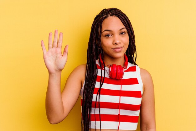 Joven afroamericana escuchando música con auriculares aislados en la pared amarilla sonriendo alegre mostrando el número cinco con los dedos