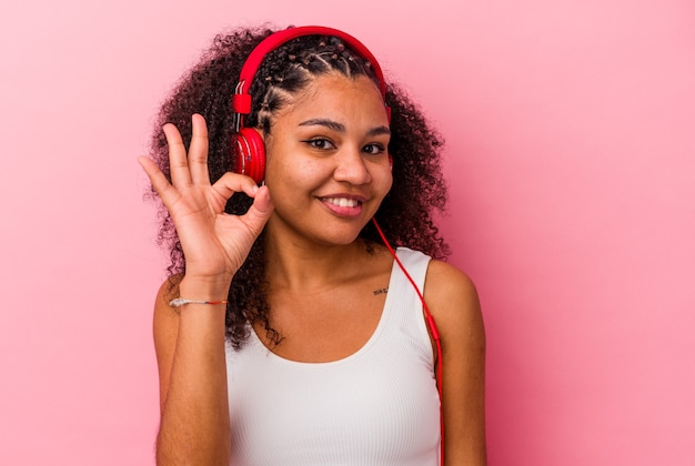 Joven afroamericana escuchando música con auriculares aislados sobre fondo rosa alegre y confiado mostrando gesto ok.