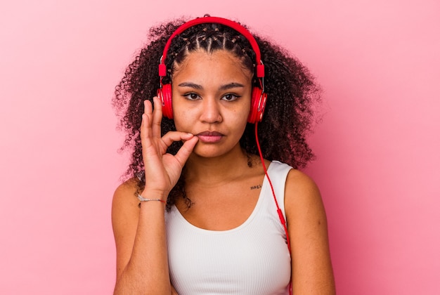 Joven afroamericana escuchando música con auriculares aislados sobre fondo rosa con los dedos en los labios guardando un secreto.