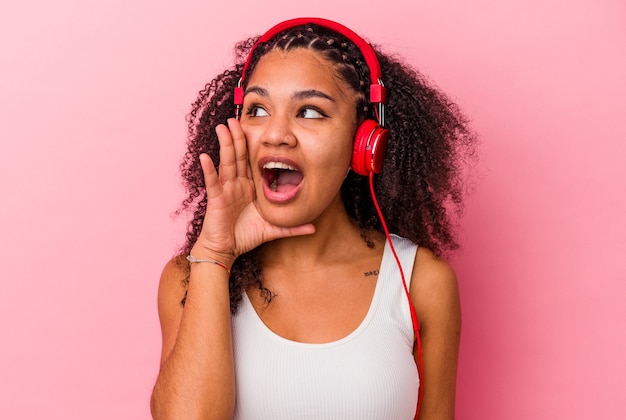 Joven afroamericana escuchando música con auriculares aislados sobre fondo rosa gritando y sosteniendo la palma cerca de la boca abierta.