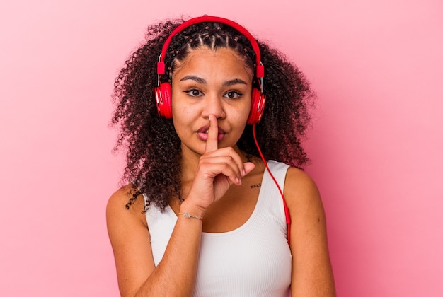 Joven afroamericana escuchando música con auriculares aislados sobre fondo rosa guardando un secreto o pidiendo silencio.