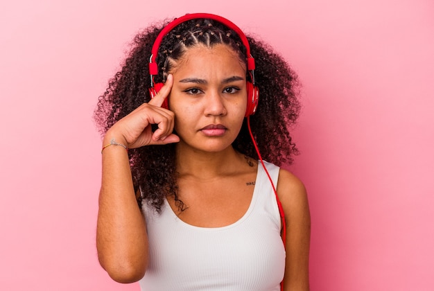 Joven afroamericana escuchando música con auriculares aislados sobre fondo rosa señalando la sien con el dedo, pensando, centrado en una tarea.