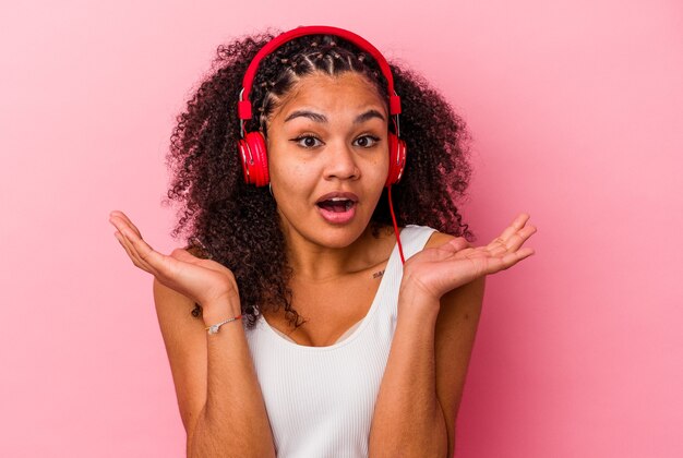 Joven afroamericana escuchando música con auriculares aislados sobre fondo rosa sorprendido y consternado.