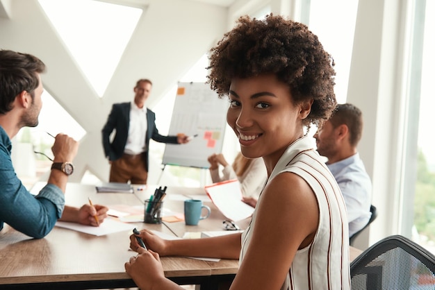 Una joven afroamericana está mirando a la cámara y sonriendo mientras su jefe está de pie cerca