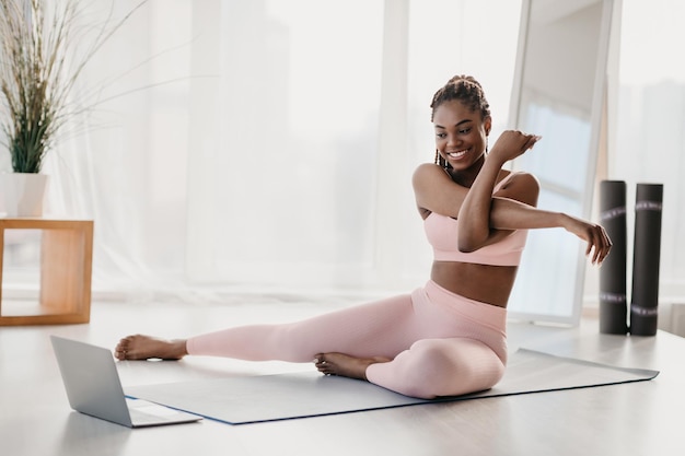 Una joven afroamericana en forma estirando su brazo en casa usando una computadora portátil viendo en línea