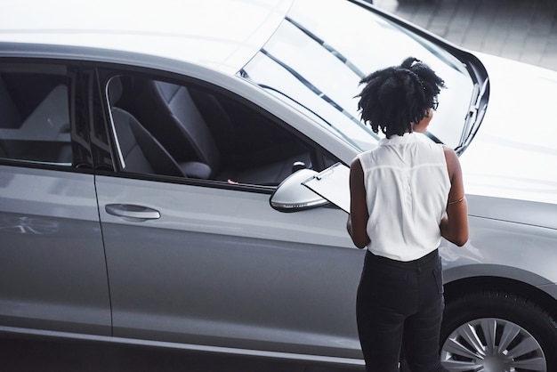 Joven afroamericana en gafas y con bloc de notas se encuentra en el salón del automóvil cerca del vehículo.