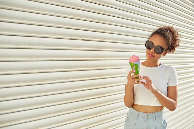 Una joven afroamericana hermosa con una camiseta blanca y jeans claros come helado contra una pared clara en un día soleado