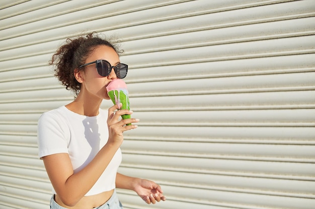 Una joven afroamericana hermosa con una camiseta blanca y jeans claros come helado contra una pared clara en un día soleado