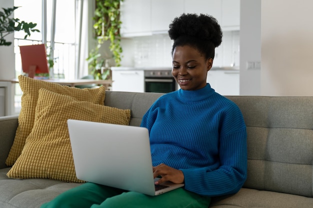 Una joven afroamericana independiente escribiendo un artículo en una laptop sentada en un sofá en un apartamento moderno