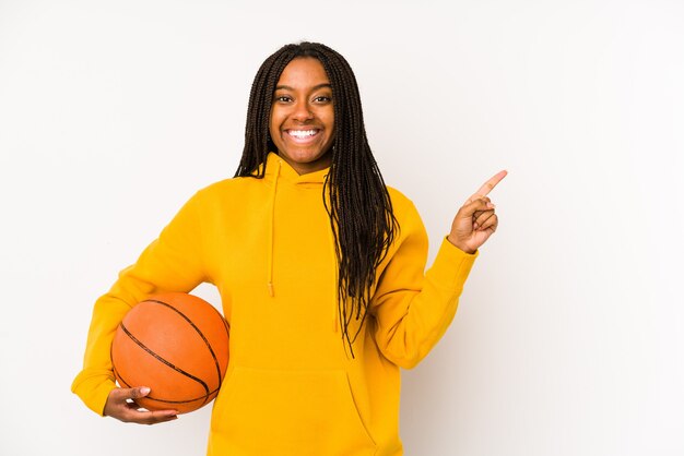 Joven afroamericana jugando baloncesto aislado sonriendo y apuntando a un lado, mostrando algo en el espacio en blanco.