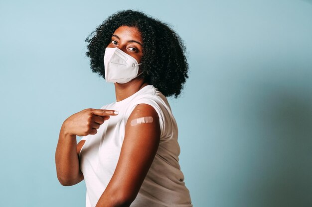 joven afroamericana con máscara apuntando a su brazo vendado para la vacunación