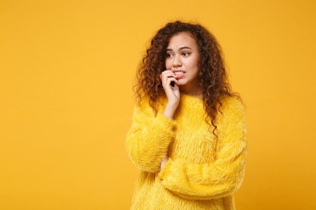 Una joven afroamericana nerviosa y preocupada con suéter de piel posando aislada en un fondo de pared naranja amarillo, retrato de estudio. Concepto de estilo de vida de las personas. Simulacros de espacio de copia. Mirando a un lado mordiéndose las uñas.