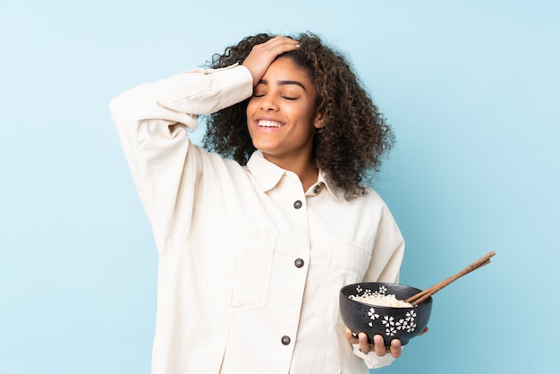 Una joven afroamericana en la pared azul se ha dado cuenta de algo y tiene la intención de la solución mientras sostiene un tazón de fideos con palillos