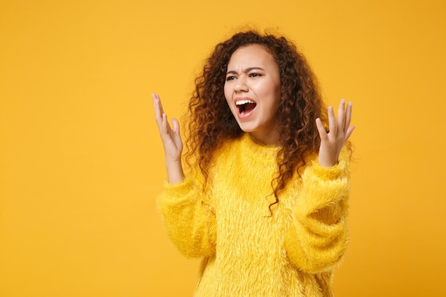 Joven afroamericana perpleja con suéter de piel posando aislada en el fondo de la pared naranja amarilla. Concepto de estilo de vida de las personas. Simulacros de espacio de copia. Extendiendo las manos, gritando jurando, mirando a un lado.