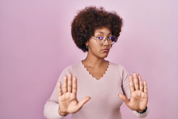 Joven afroamericana de pie sobre un fondo rosado alejándose de las manos mostrando rechazo y negación con expresión de miedo y repugnante. parada y prohibido.