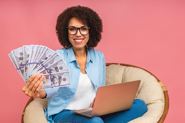 Joven afroamericana positiva negra positiva con el pelo rizado usando una laptop y sonriendo aislada sobre un fondo rosado Sosteniendo billetes de dinero