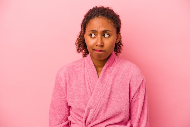 Joven afroamericana que usa una bata de baño aislada en un fondo rosa confundida se siente dudosa e insegura