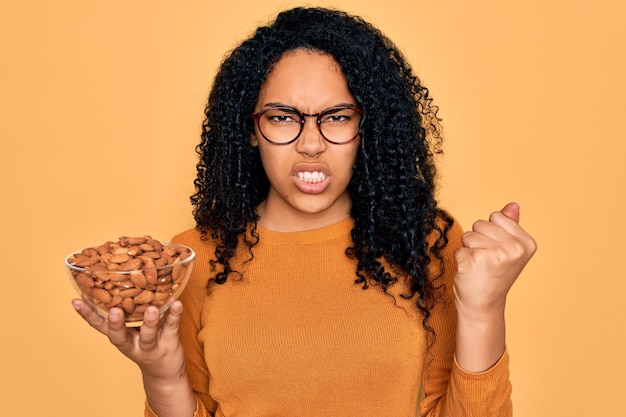 Joven afroamericana rizada sosteniendo un tazón con almendras saludables sobre fondo amarillo molesta y frustrada gritando con ira loca y gritando con el concepto de ira de mano levantada