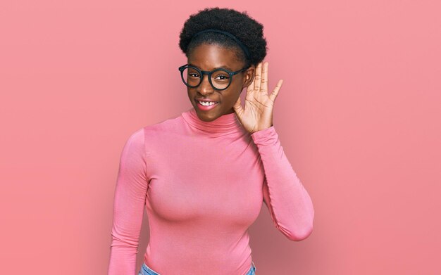 Joven afroamericana con ropa informal y anteojos sonriendo con la mano sobre la oreja escuchando rumores o chismes sobre el concepto de sordera