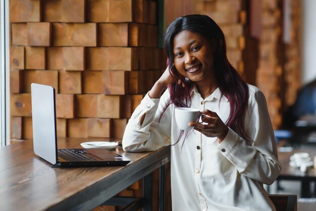 Joven afroamericana sentada en el restaurante y escribiendo en su computadora portátil Chica guapa trabajando en la computadora en el café