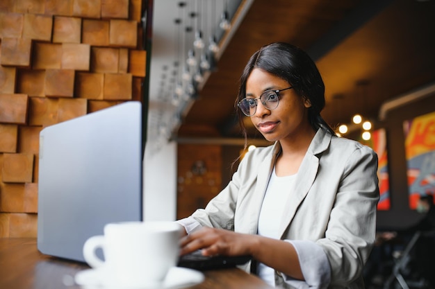 Joven afroamericana sentada en un restaurante y escribiendo en su laptop. Linda chica trabajando en la computadora en el café.