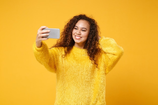 Una joven afroamericana sonriente con suéter de piel posando aislada en un fondo de pared naranja amarillo, retrato de estudio. Concepto de estilo de vida de las personas. Simulacros de espacio de copia. Haciendo autofoto en el teléfono móvil.