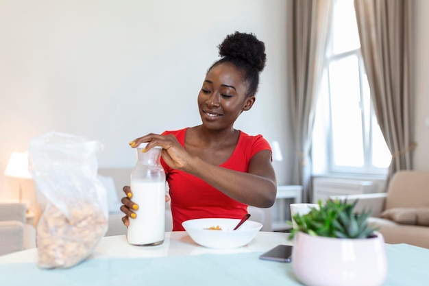 Una joven afroamericana sonriente vierte copos de maíz en un plato con leche La niña tiene un desayuno saludable en una casa elegante y acogedora por la mañana mientras revisa su correo electrónico en una laptop