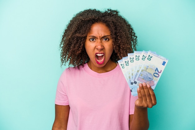 Joven afroamericana sosteniendo billetes en euros aislado sobre fondo azul gritando muy enojado y agresivo.