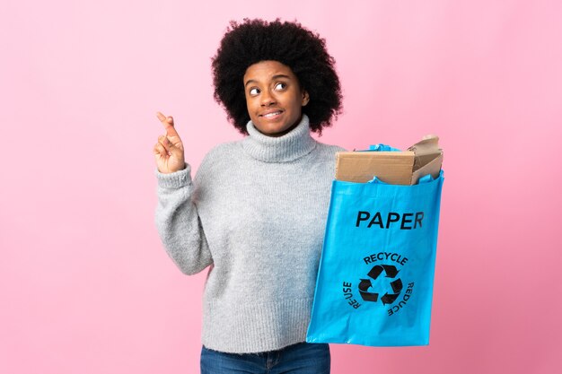 Joven afroamericana sosteniendo una bolsa de reciclaje con los dedos cruzando y deseando lo mejor
