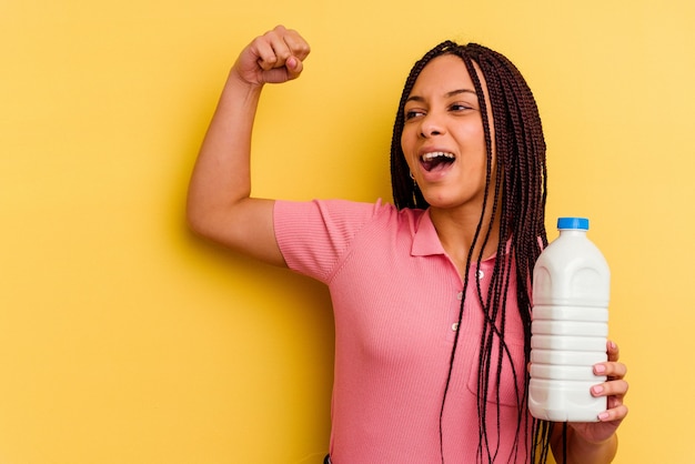 Joven afroamericana sosteniendo una botella de leche aislada en amarillo levantando el puño después de una victoria, concepto ganador.