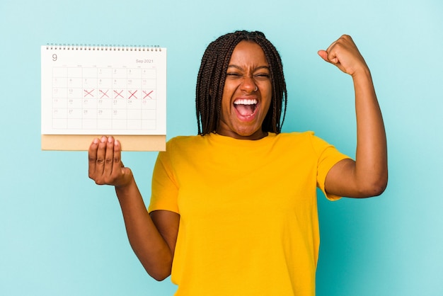 Joven afroamericana sosteniendo un calendario aislado sobre fondo azul levantando el puño después de una victoria, concepto ganador.