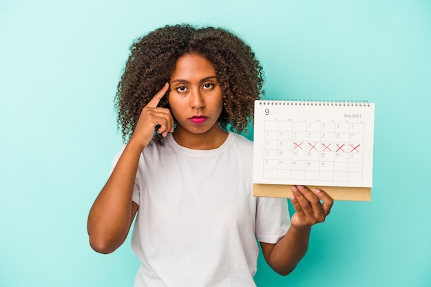 Joven afroamericana sosteniendo un calendario aislado sobre fondo azul señalando el templo con el dedo, pensando, centrado en una tarea.