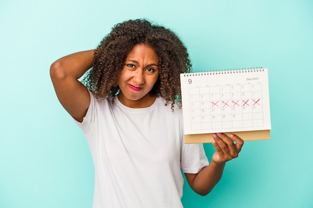 Joven afroamericana sosteniendo un calendario aislado sobre fondo azul tocando la parte posterior de la cabeza, pensando y tomando una decisión.