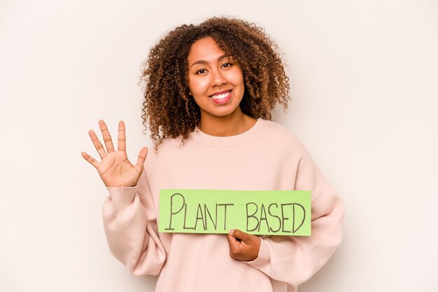 Joven afroamericana sosteniendo un cartel a base de plantas aislado de fondo blanco sonriendo alegre mostrando el número cinco con los dedos