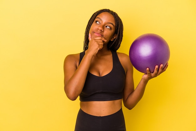 Joven afroamericana sosteniendo una pelota de pilates aislada sobre fondo amarillo mirando hacia los lados con expresión dudosa y escéptica.