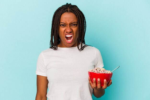 Joven afroamericana sosteniendo un plato de cereales aislado sobre fondo azul gritando muy enojado y agresivo.