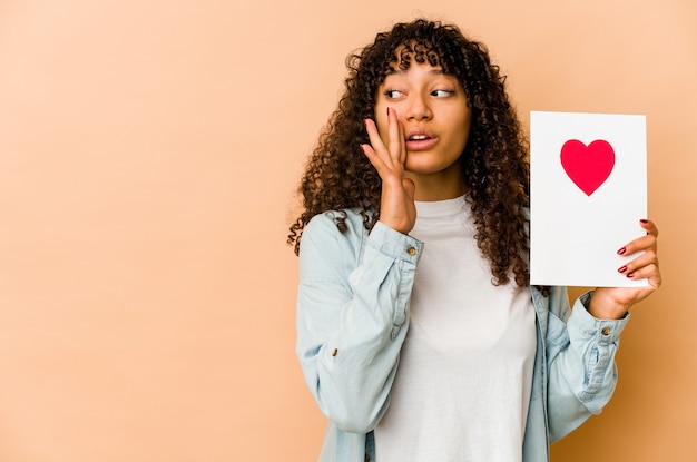 Joven afroamericana sosteniendo una tarjeta del día de San Valentín está diciendo una noticia secreta de frenado caliente y mirando a un lado