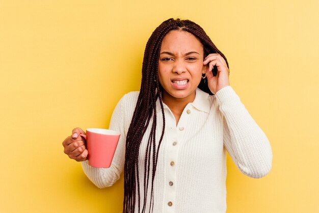 Joven afroamericana sosteniendo una taza aislada cubriendo las orejas con las manos.