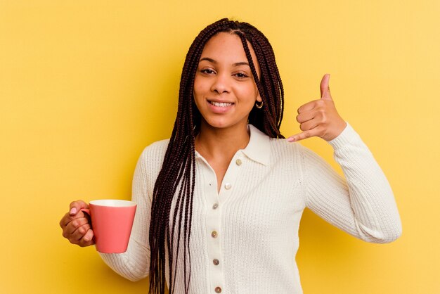 Joven afroamericana sosteniendo una taza aislada mostrando un gesto de llamada de teléfono móvil con los dedos.