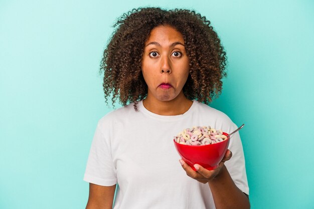 Joven afroamericana sosteniendo un tazón de cereales aislado sobre fondo azul se encoge de hombros y abre los ojos confundidos.