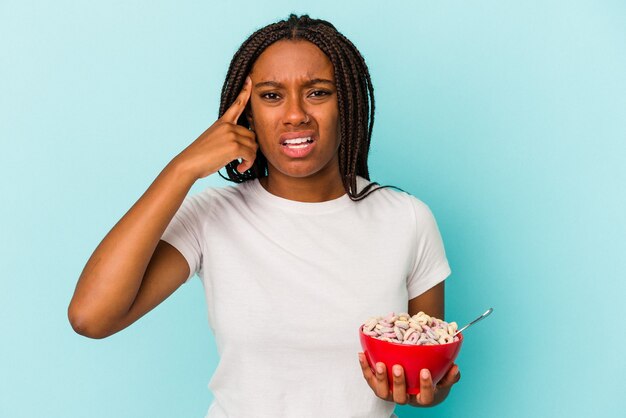 Joven afroamericana sosteniendo un tazón de cereales aislado sobre fondo azul mostrando un gesto de decepción con el dedo índice.