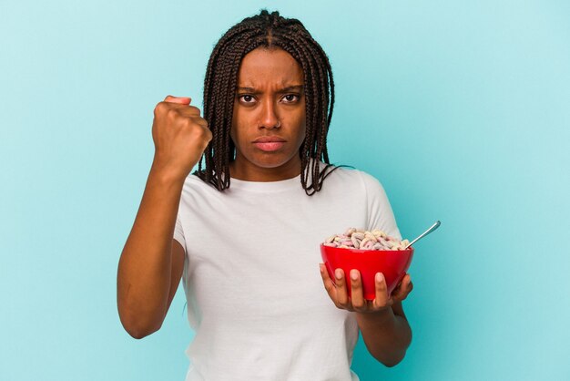 Joven afroamericana sosteniendo un tazón de cereales aislado sobre fondo azul mostrando el puño a la cámara, expresión facial agresiva.