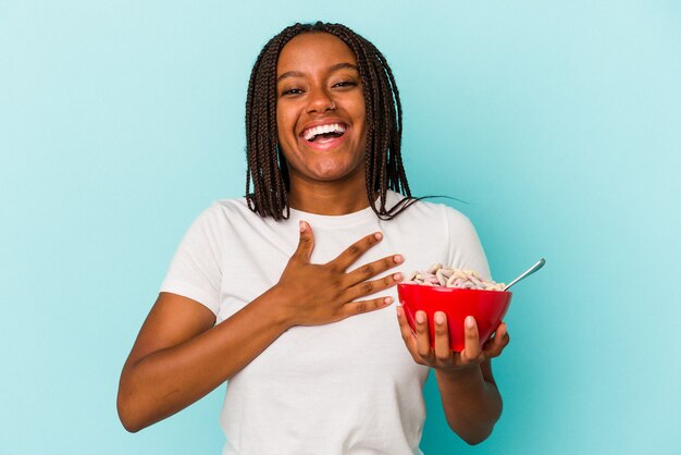 Joven afroamericana sosteniendo un tazón de cereales aislado sobre fondo azul se ríe a carcajadas manteniendo la mano en el pecho.