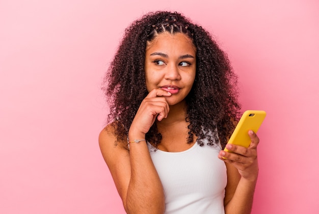 Joven afroamericana sosteniendo un teléfono móvil aislado una pared rosa relajada pensando en algo mirando un espacio de copia
