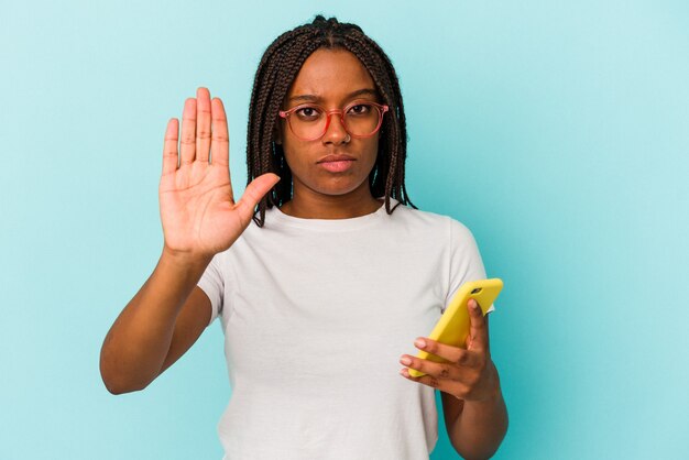 Joven afroamericana sosteniendo un teléfono móvil aislado sobre fondo azul de pie con la mano extendida mostrando la señal de stop, impidiéndote.