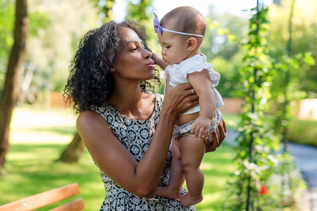 Joven afroamericana y su hija caminando en el parque de verano. Madre negra jugando con su bebé al aire libre.