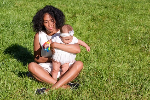 Joven afroamericana y su hija sentada sobre la hierba verde en el parque de verano.