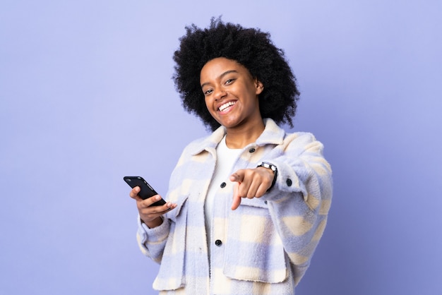 Joven afroamericana con teléfono móvil aislado sobre fondo púrpura le señala con el dedo con una expresión de confianza